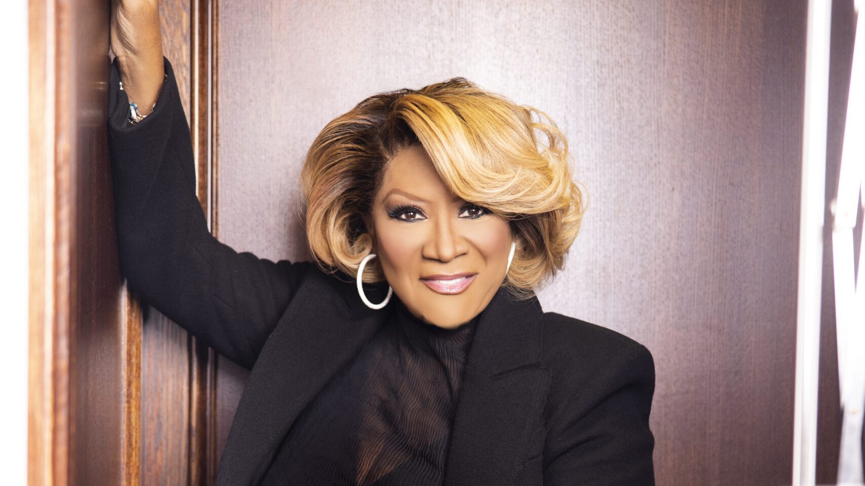 Patti LaBelle, smiling, wearing an elegant black outfit leans against a door frame.